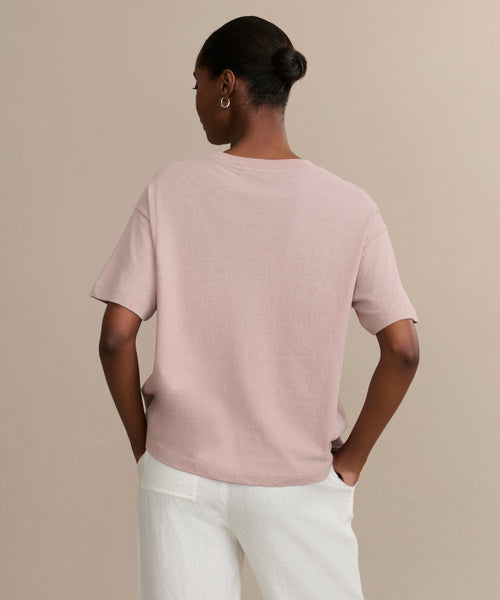 A person stands with hands on hips, wearing the Vintage Pocket Tee in light pink and white pants. Their hair is styled in a neat bun with hoop earrings, creating an effortless casual look against a plain beige background.