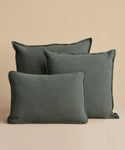 Three Adler Lumbar Pillows in a dark grayish-green linen blend are arranged against a beige background, with two standing upright at the back and one lying flat in front.