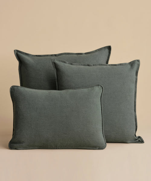 Three Adler Lumbar Pillows in a dark grayish-green linen blend are arranged against a beige background, with two standing upright at the back and one lying flat in front.