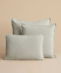 Three Adler Lumbar Pillows in light gray linen blend, inspired by Belgian design and varying in size, are arranged staggered with the smallest in front against a plain beige background.
