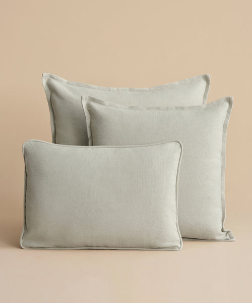 Three Adler Lumbar Pillows in light gray linen blend, inspired by Belgian design and varying in size, are arranged staggered with the smallest in front against a plain beige background.