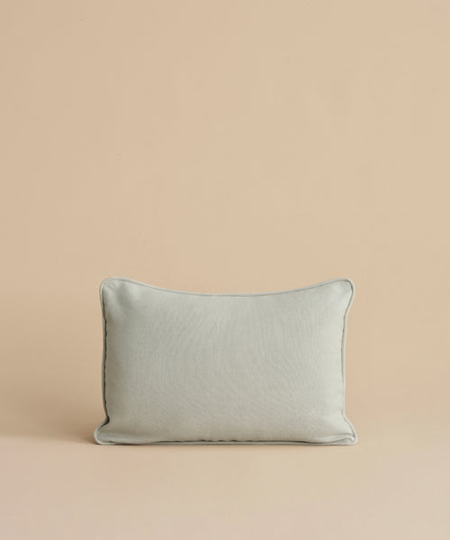 The Adler Lumbar Pillow, featuring a light gray linen blend with dark piping, is displayed upright against a plain beige background.