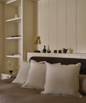 A cozy, neutral bedroom with three light beige Adler Pillows on a brown bed, built-in shelves holding books and vases, a small black lamp, and minimalist black decor displayed on the headboard shelf.