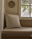 A cozy corner bench with a light plaid cushion and the Adler Pillow sits by a window with sunlight streaming in and a round textured wall decor above, while green vines are visible outside.