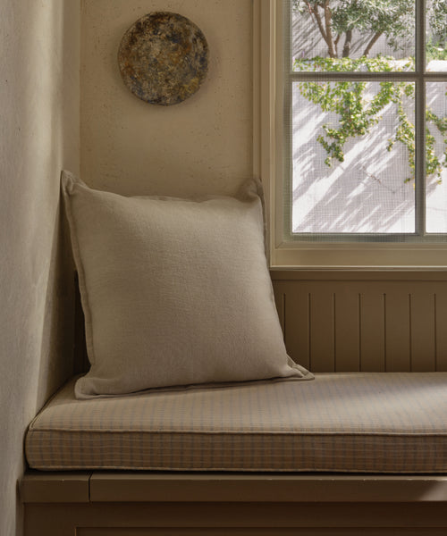A cozy corner bench with a light plaid cushion and the Adler Pillow sits by a window with sunlight streaming in and a round textured wall decor above, while green vines are visible outside.
