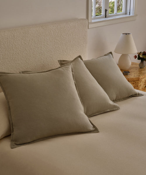 Three large beige Adler Pillows are arranged neatly on a bed with a matching linen blend comforter. A nightstand with a lamp and flower arrangement sits next to the bed, beneath a window with white trim.