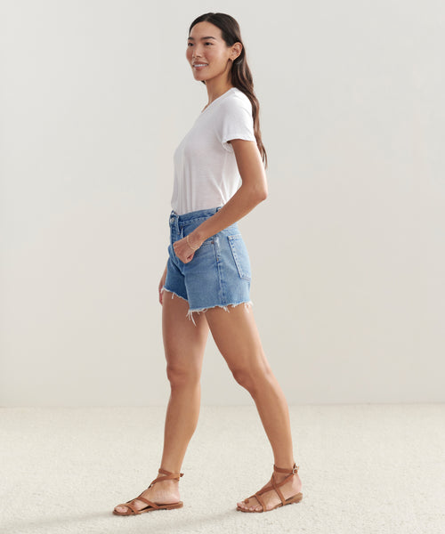 A woman stands sideways, smiling, in a white t-shirt, Parker Long Short by AGOLDE denim vintage fit shorts, and brown sandals. She poses indoors against a plain light-colored background.