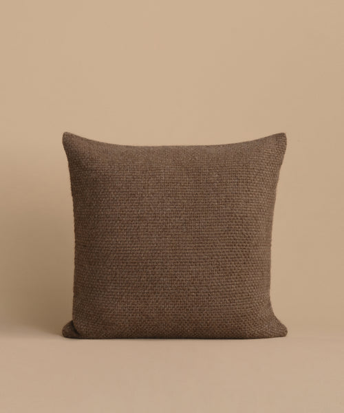 The Alpaca Basketweave Pillow, a handmade square brown cushion from Peru, is displayed upright against a beige background.