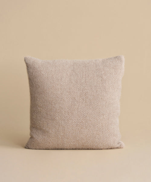 The Alpaca Basketweave Pillow, featuring a textured woven design and handmade in Peru, stands upright against a plain, neutral beige background.