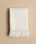 The Alpaca Basketweave Throw, handwoven in Peru, features an off-white color with fringe and is neatly folded on a neutral beige background.
