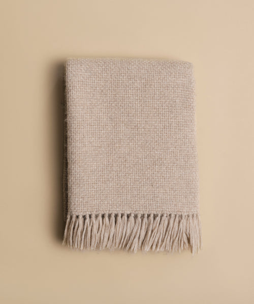 The Alpaca Basketweave Throw, in beige with fringed edges, is folded on a plain, light tan background, highlighting its super-soft, handwoven Peruvian texture.