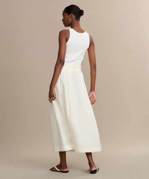 A woman faces away, wearing the Annabel Skirt— a high-waisted cream linen piece—paired with a white sleeveless top and flat sandals. Her hair is styled in a neat bun against a neutral background.