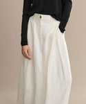 Someone wears the Annabel Skirt, a vintage-inspired, high-waisted off-white linen piece with pleats, styled with a black long-sleeve top. Only the lower body is shown against a plain beige background.