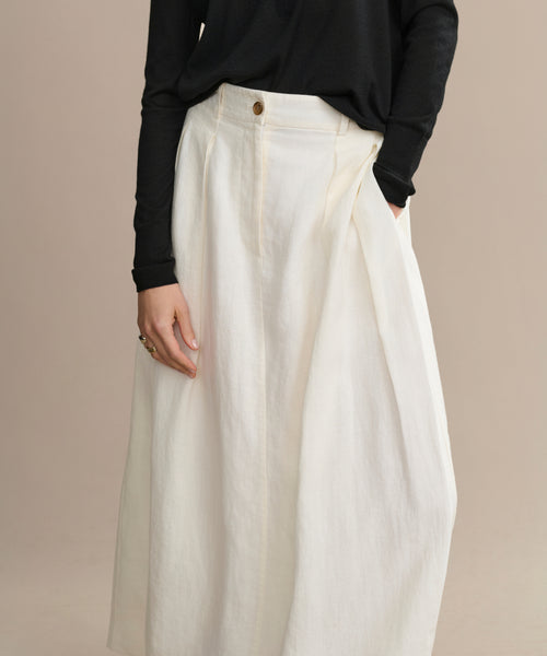 Someone wears the Annabel Skirt, a vintage-inspired, high-waisted off-white linen piece with pleats, styled with a black long-sleeve top. Only the lower body is shown against a plain beige background.
