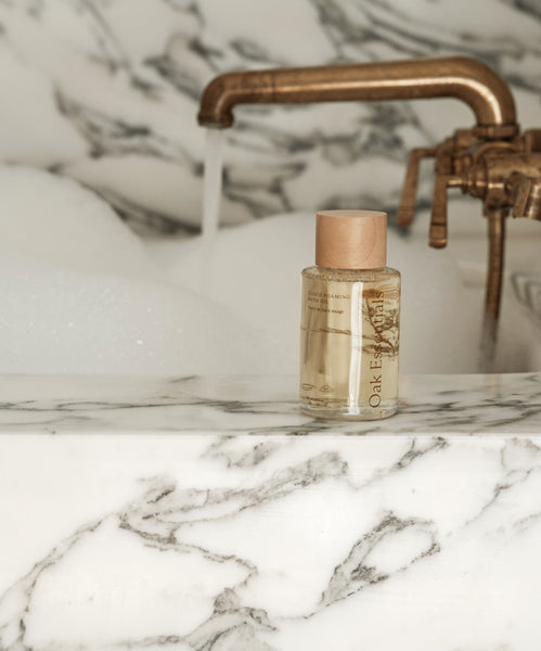 A bottle of bath oil from the Aromatherapy Bath Set rests on a white marble tubs edge, as water pours from a brass faucet and bubbles fill the background.