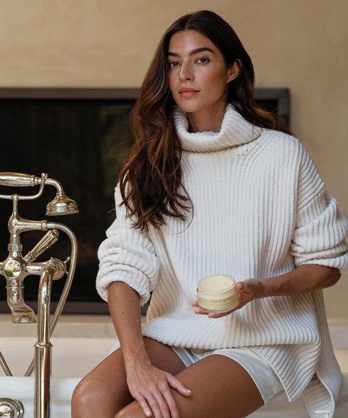 A woman in a white turtleneck sweater with long brown hair sits by a bathtub, holding an Aromatherapy Bath Set. She has a calm expression, and the room is softly lit with natural light.