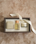A beige gift box holds the Aromatherapy Bath Set—ideal for an at-home spa—with a soap bar, hydrating body balm, and body oil with a wooden cap, topped with a white satin ribbon.