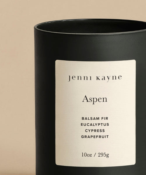 The Aspen Glass Candle is a black glass container with a white jenni kayne Aspen label. Scented with Balsam Fir, Eucalyptus, Cypress, and Grapefruit. 10oz/295g natural wax blend, hand-poured in LA, shown on a beige background.