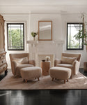Two Aspen Chairs in soft Sherpa fabric with matching ottomans rest on a light rug by a white fireplace. Black-framed windows and a wooden side table with flowers enhance this inviting, mid-century modern living room.