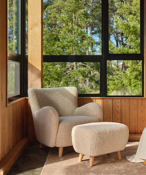 The Aspen Chair with Sherpa upholstery and its matching ottoman rest on a tan rug in a sunlit, mid-century modern room featuring wood-paneled walls and large windows that overlook green trees.