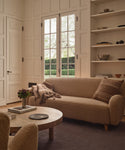 A cozy living room with an Aspen Sofa in Oatmeal Sherpa, a round wood coffee table with flowers, open shelves decorated with accents, and large windows for a bright, mid-century modern atmosphere.