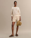 A woman stands against a beige background wearing the Avery Crewneck, a lightweight knit striped sweater, paired with beige shorts, brown sandals, and holding a woven sun hat.