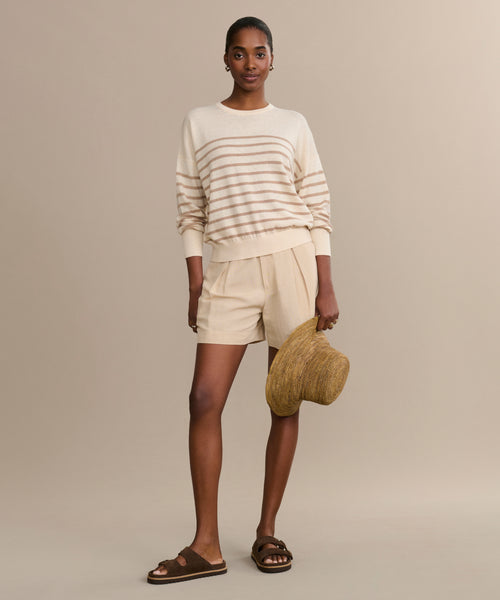 A woman stands against a beige background wearing the Avery Crewneck, a lightweight knit striped sweater, paired with beige shorts, brown sandals, and holding a woven sun hat.