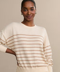 A woman with dark skin and hair pulled back smiles at the camera, wearing the Avery Crewneck sweater and gold hoop earrings, standing against a plain beige background.