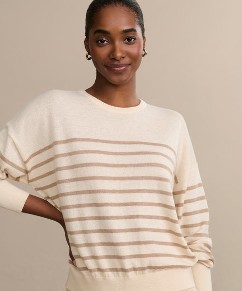 A woman with dark skin and hair pulled back smiles at the camera, wearing the Avery Crewneck sweater and gold hoop earrings, standing against a plain beige background.