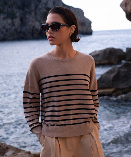 A woman with short dark hair and black sunglasses stands by the rocky seashore in beige pants, wearing the Avery Crewneck, creating a relaxed coastal look with cliffs and water behind her.