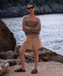 Wearing the Avery Crewneck striped sweater, sunglasses, tan shorts, and brown sandals, a person stands on rocky ground by the sea, exuding a relaxed coastal vibe with cliffs and water in the background.
