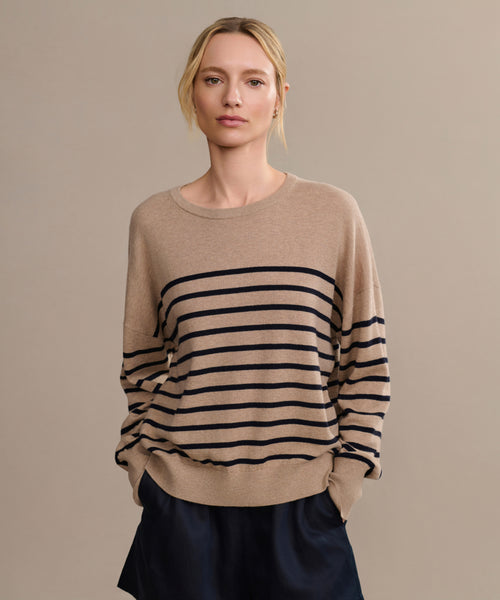A woman stands against a beige background, wearing the Avery Crewneck—a loose cotton-merino sweater with black stripes—and dark shorts, her hands in her pockets and a neutral expression.