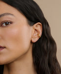 Close-up of a woman’s ear wearing the Bagel Ear Cuff—a small, gold accessory ideal for everyday style. She has smooth, medium-toned skin and long, wavy dark hair against a beige background.