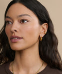 A woman with long, wavy dark hair wears a Bagel Ear Cuff and gold hoop earrings, paired with a gold necklace. She poses in natural makeup and a brown top against a neutral beige background, showcasing an everyday modern jewelry look.