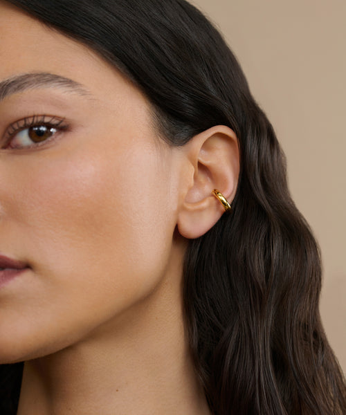 Close-up of a woman’s ear wearing the Bagel Ear Cuff—a small, gold accessory ideal for everyday style. She has smooth, medium-toned skin and long, wavy dark hair against a beige background.