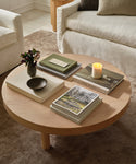 The Bay Coffee Table, crafted from knotty alder, displays stacked books, a lit candle, a small floral vase, and a decorative bowl in a cozy living room with neutral sofas and a brown rug.
