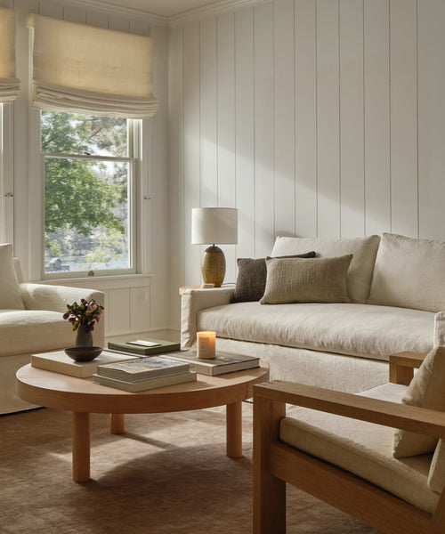 A cozy living room features cream sofas and a handcrafted Bay Coffee Table adorned with books, a candle, and flowers. Sunlight filters through windows with light Roman shades, creating a warm and inviting atmosphere.