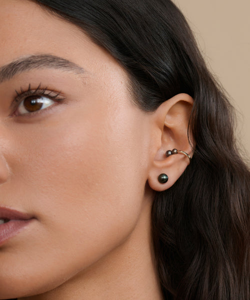 Close-up of a woman's left ear with two black studs, a silver cartilage hoop, and the Loren Pearl Ear Cuff by Jenni Kayne Jewelry. She has long dark hair, smooth skin, and is set against a neutral beige background.
