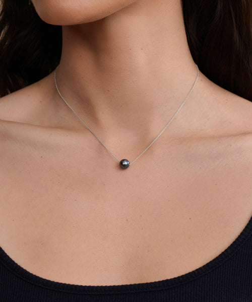 A close-up of a woman's neck wearing the Philo Pearl Necklace, featuring a single round black pearl pendant, paired with a black ribbed top.