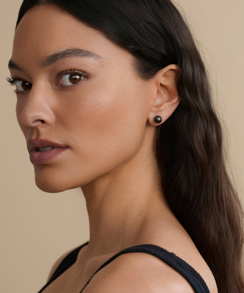 A woman with long, wavy dark hair poses in a black top, wearing Medium Loren Pearl Studs from Modern Elegance Jewelry. She faces the camera at a slight angle against a neutral beige background.