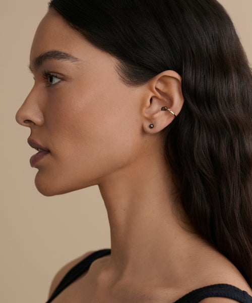 A woman with long dark hair is shown in profile against a beige background, wearing a black top and small Loren Pearl Studs by Jenni Kayne Jewelry.