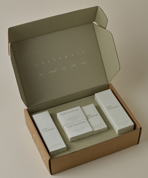 An open cardboard box displays The Body Routine by Oak Essentials, with skincare and body care products arranged neatly inside. The lid reads, CELEBRATE the ritual of self care—a true luxury beauty experience.