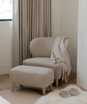 A Brentwood Chair with matching ottoman, upholstered in Belgian wool, sits near a window with sheer curtains on a light wood floor. A beige throw blanket is draped over the chair, and a fuzzy rug lies nearby.