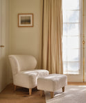 A cozy, cream-colored armchair with the Brentwood Ottoman rests on a wooden floor by beige curtains and a sunlit window. A framed artwork above complements the minimalist, serene decor.