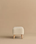 The Brentwood Ottoman is a small, square piece in cream wool boucle with textured fabric and four light wooden legs, shown on a plain beige background.