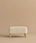 The Brentwood Ottoman is a compact, rectangular piece with off-white wool boucle upholstery and four short wooden legs, shown against a plain beige background.