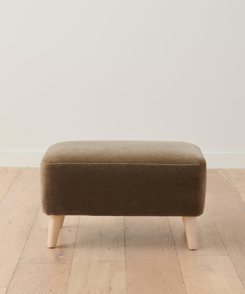 The Brentwood Ottoman is a rectangular, brown upholstered luxury ottoman with light wooden legs, positioned on a pale wood floor in front of a plain white wall.