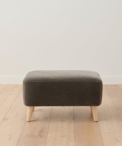 The Brentwood Ottoman, a compact rectangular piece in dark gray with light wooden legs, sits on a light wood floor by a white wall, adding stylish, modern upholstered accent seating to any space.
