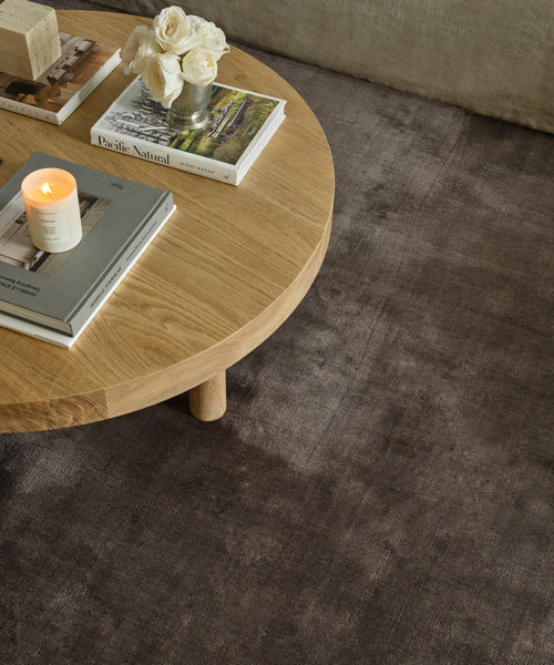 A round wooden coffee table with books, a lit candle, and a small vase of white roses sits atop the luxurious Bristol Rug, creating a cozy living room setting.