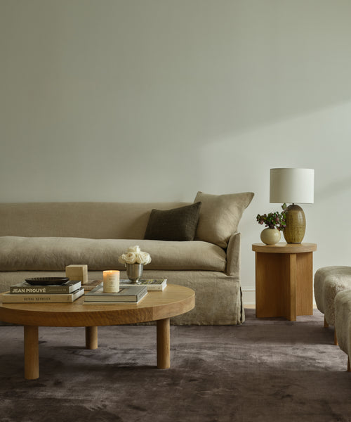 A minimalist living room with a beige sofa, brown cushion, round wooden coffee table with books, flowers, and a candle, plus a side table with lamp and vase—all set on the Bristol Rug for added warmth and character.